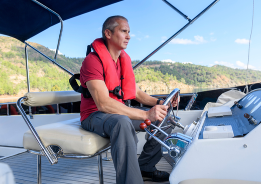 A man in a life jacket sits at the wheel of a boat.