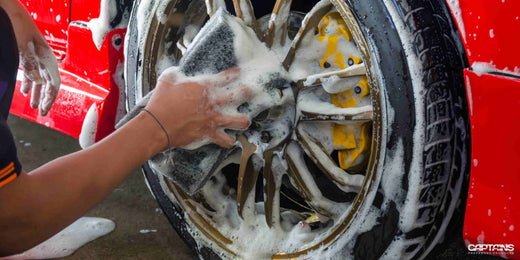 A person washing the wheels of a red sports car with a foamy sponge.