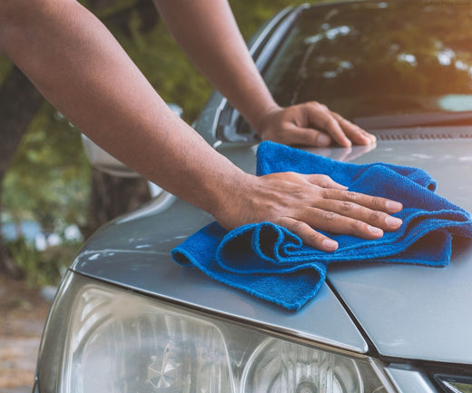 A person drying a silver car hood with a blue microfiber cloth.