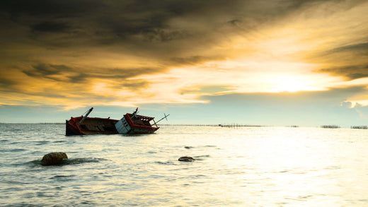 A capsized boat sitting halfway out of the water against a yellow sunset.