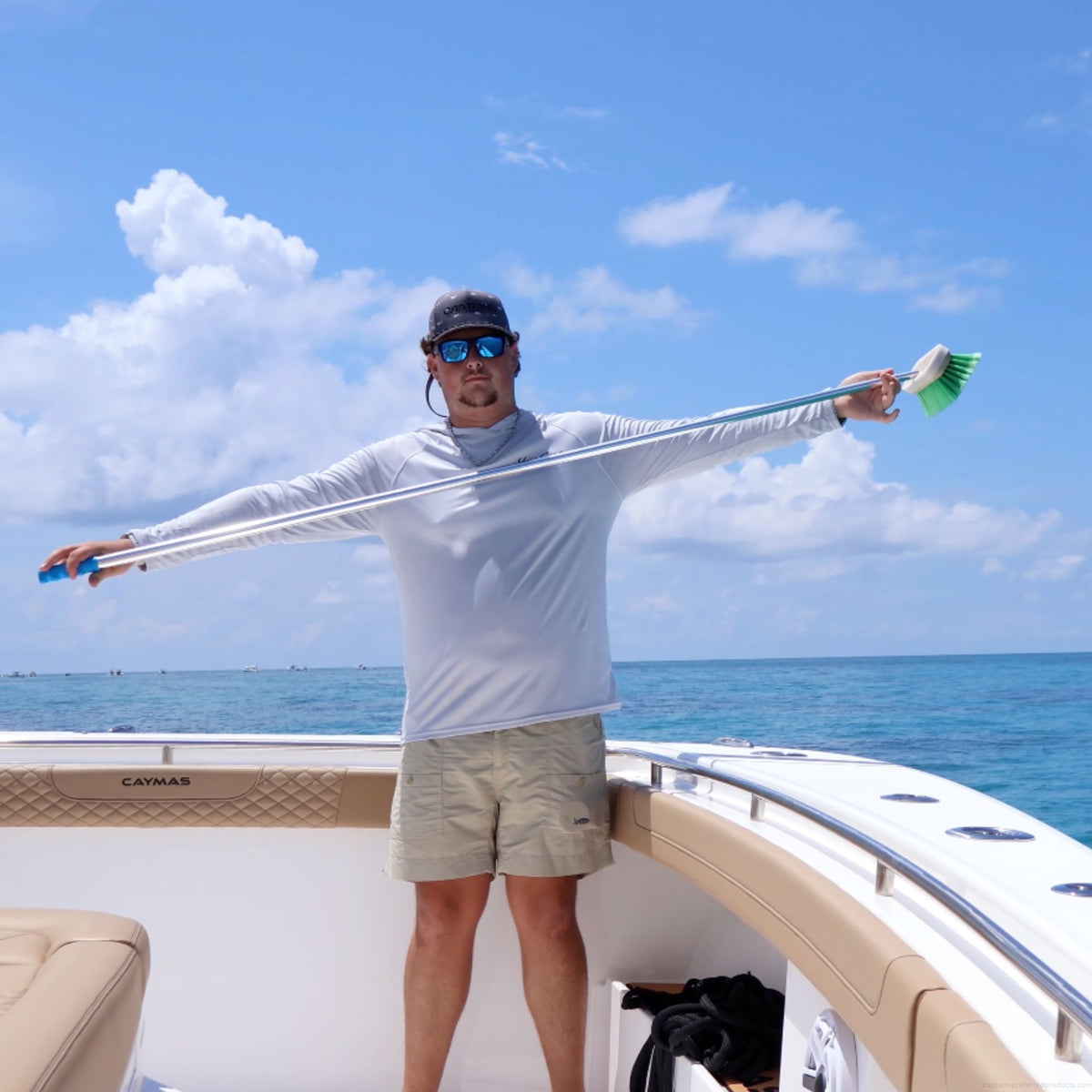 A man wearing a white long-sleeve fishing shirt and beige shorts stands on the deck of a boat, stretching the long handle of a boat brush with green bristles out to both sides against a blue ocean and sky background.