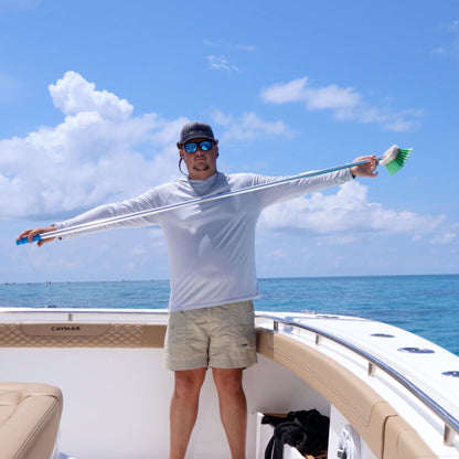 A man wearing a white long-sleeve fishing shirt and beige shorts stands on the deck of a boat, stretching the long handle of a boat brush with green bristles out to both sides against a blue ocean and sky background.