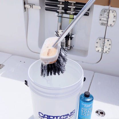 A black hard bristled boat brush is dunked into a bucket of soapy water to clean a boat deck.