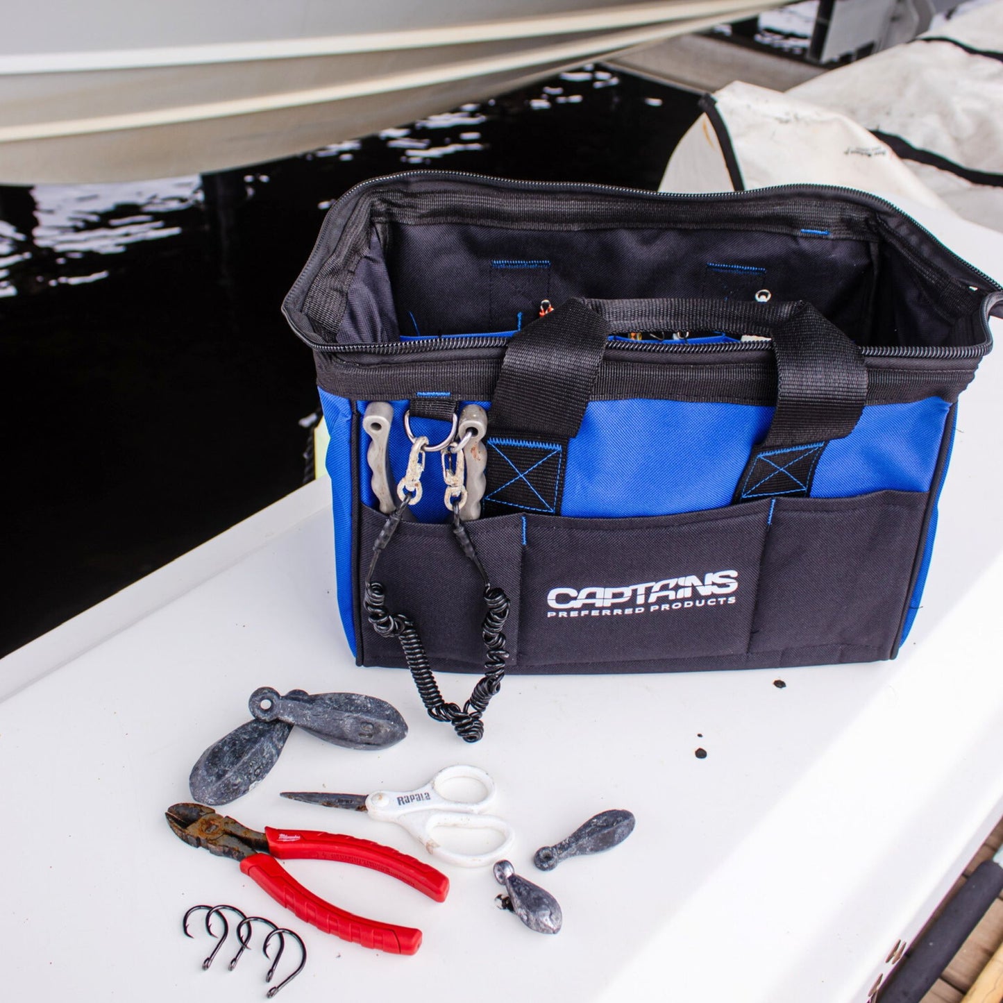 A fishing tool bag sits on the edge of a dock with tools and fishing gear around it.
