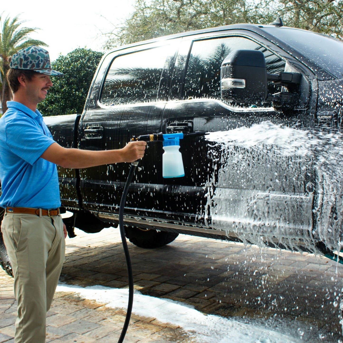 Man washing a black truck with a foam cannon and hose on a sunny day.